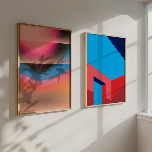 Two abstract art pieces in gold frames on a white wall with sunlight casting shadows.