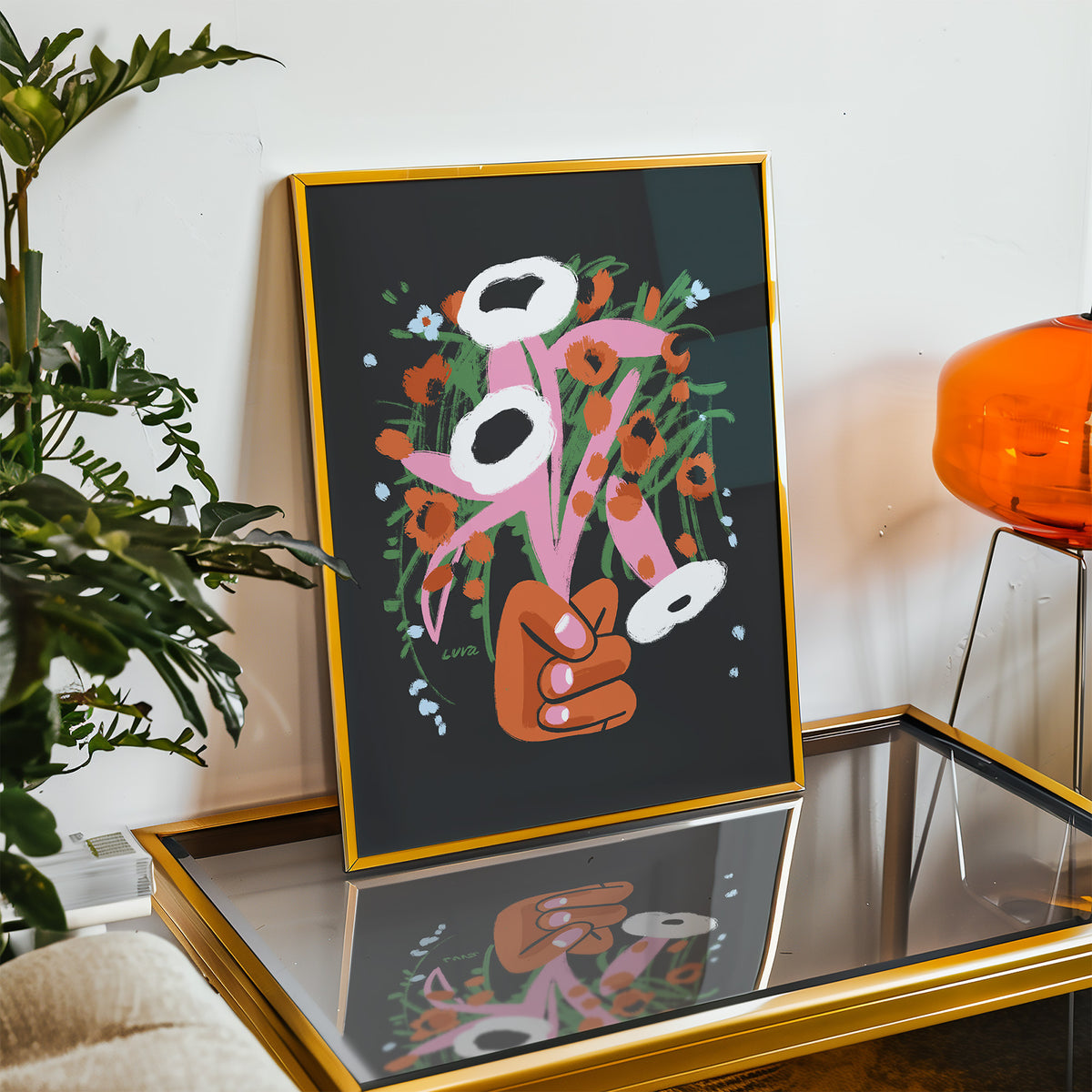 Framed artwork of a hand holding flowers on a reflective surface with a plant and lamp in the background.