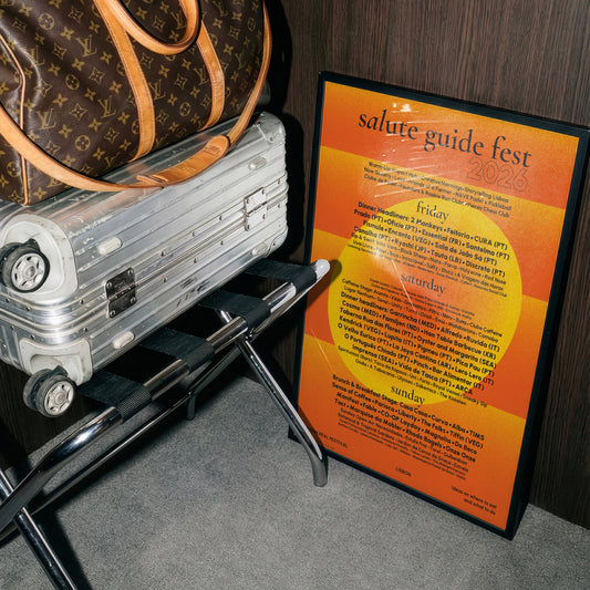 A framed poster reading 'salute guide fest 2026' with orange and yellow gradient text, leaning on a black metal stand next to a silver suitcase and a brown bag.