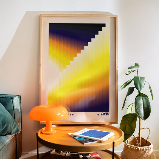 Framed abstract art piece on a wall above a round wooden table with a lamp and books.