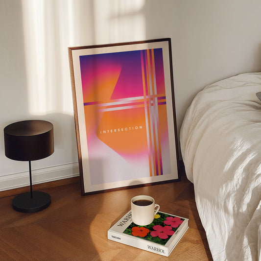 Framed abstract art on a wooden floor next to a bed with a coffee cup and book.
