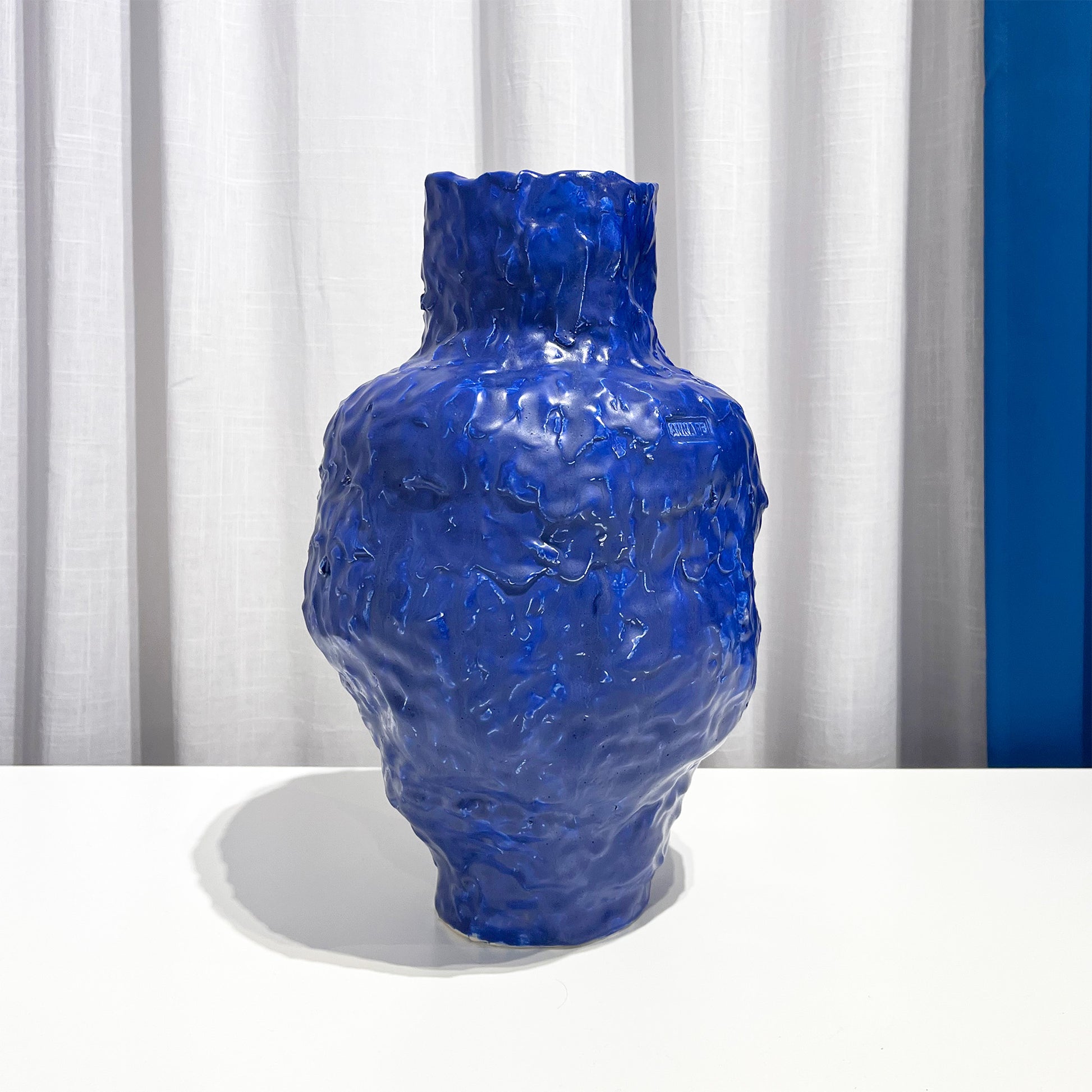 Blue textured vase on a white surface with a white curtain background by Anna De at Levö