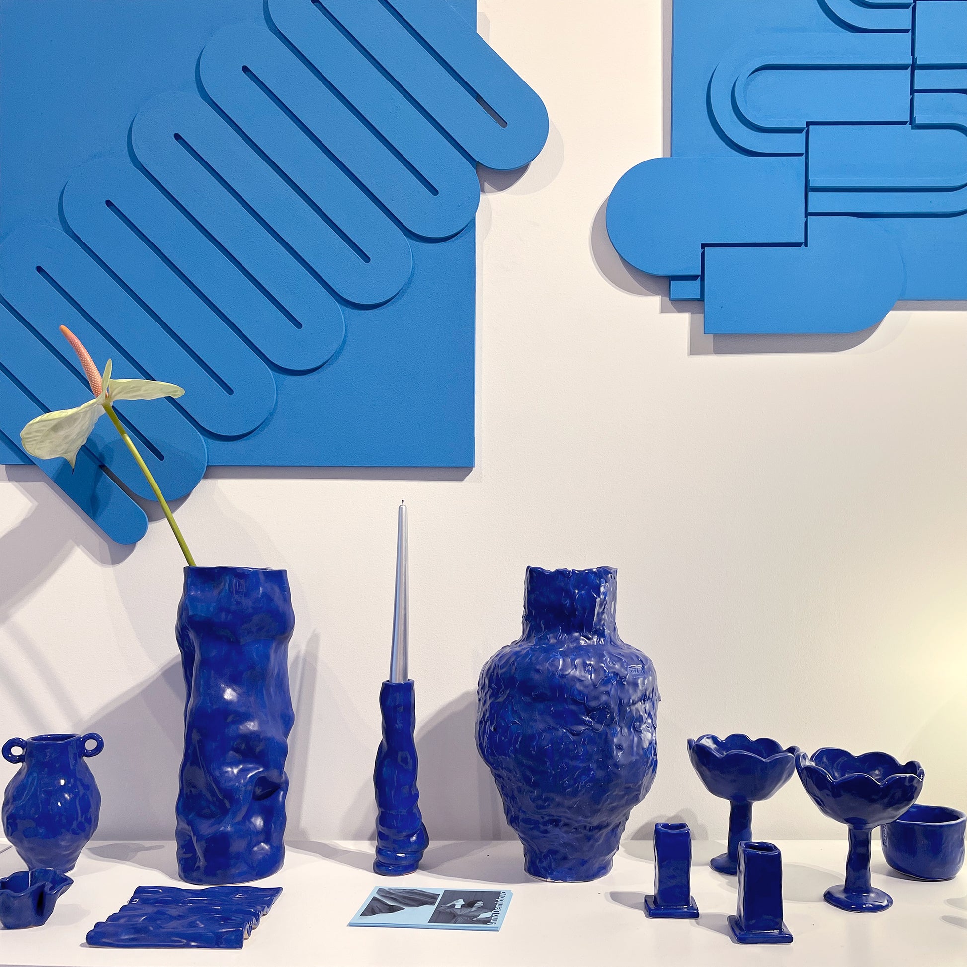 Collection of blue ceramic vases and decorative items against a white wall with blue abstract Levö