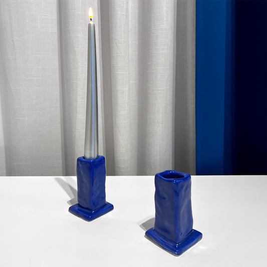 Two blue candle holders with a tall gray candle against a white curtain background