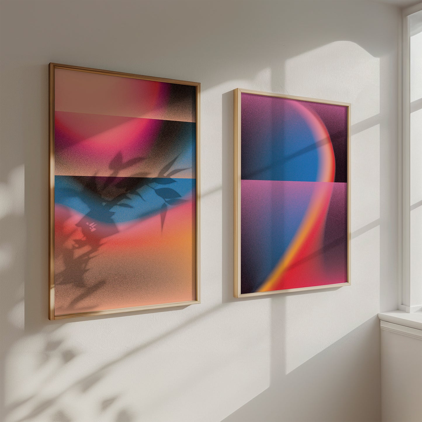 Two abstract art pieces in wooden frames on a white wall with sunlight casting shadows.