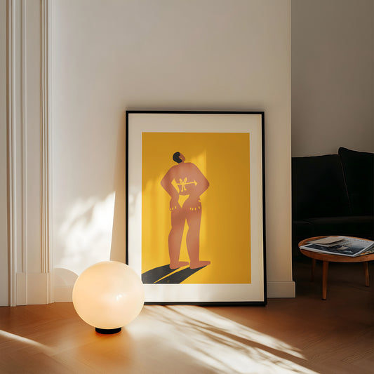 Framed artwork of a person on a yellow background in a room with a lamp and table.