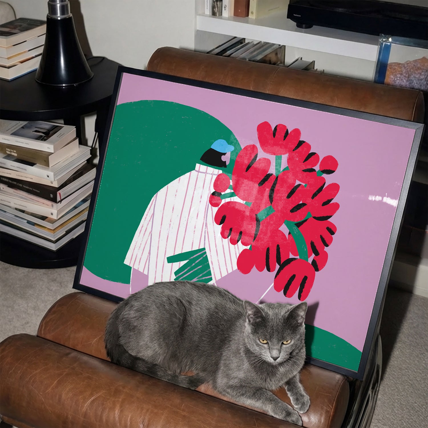 Gray cat sitting on a brown armchair next to a framed pink poster by Levö with a boy and red flowers.