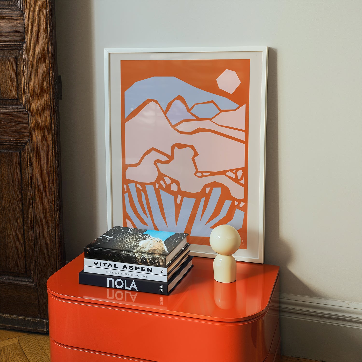 Framed abstract artwork on a wall above an orange side table with books and a decorative object.