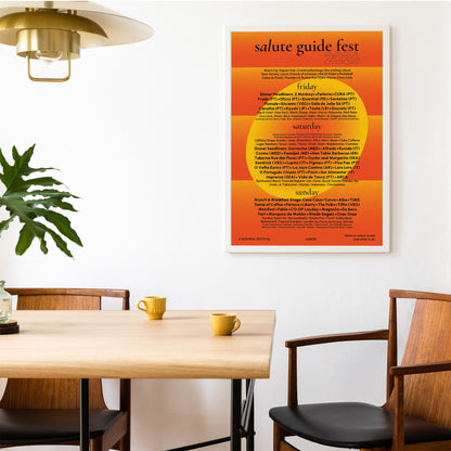 A framed poster titled 'salute guide fest 2026' with an orange and yellow schedule layout, displayed on a white wall above a wooden dining table with chairs and yellow mugs in a modern interior setting.